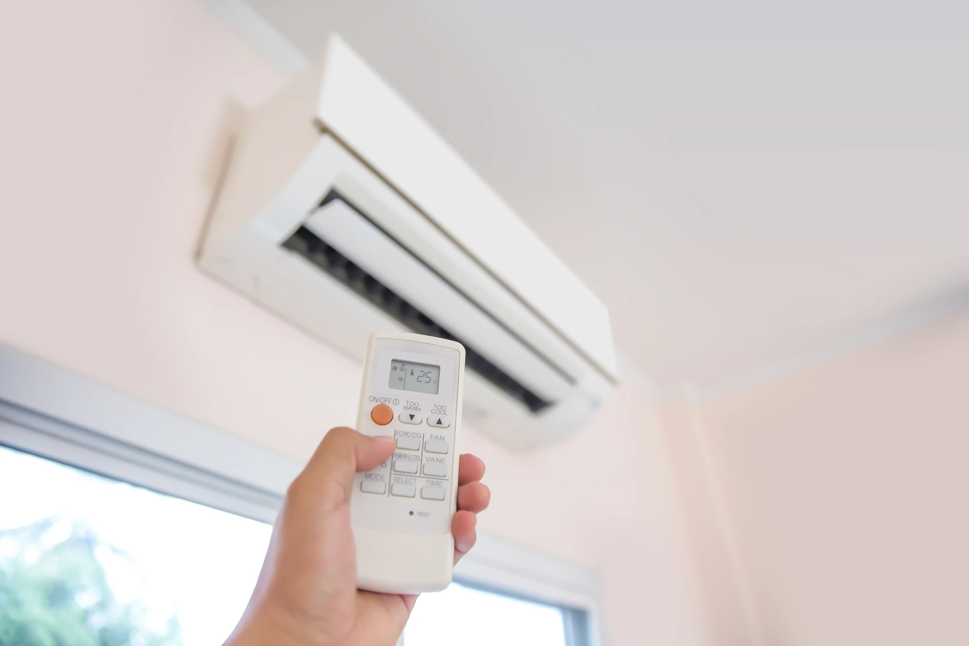 Main tenant une t&eacute;l&eacute;commande blanche dirig&eacute;e vers une unit&eacute; de climatisation murale split install&eacute;e au plafond d'un int&eacute;rieur r&eacute;sidentiel