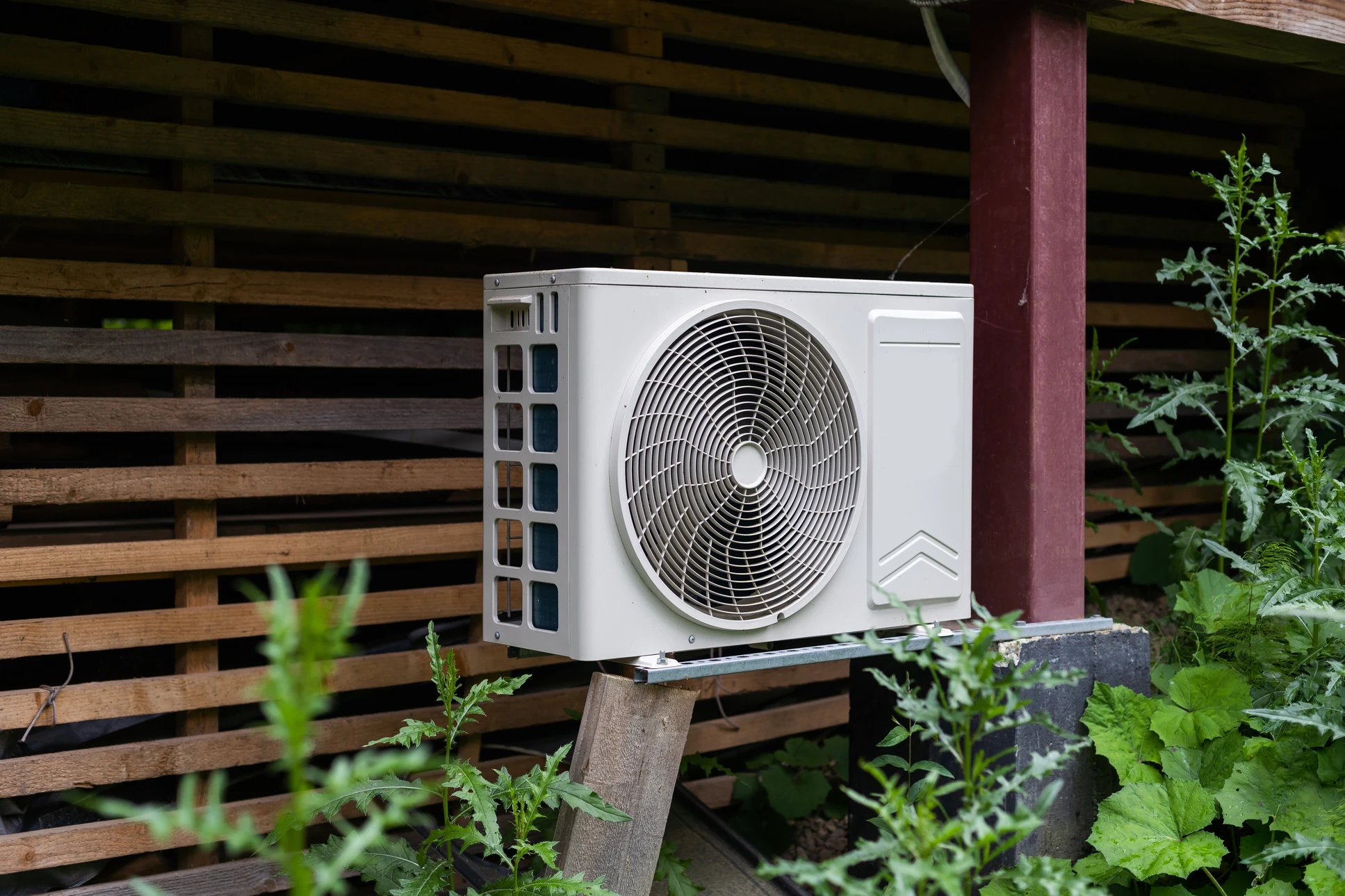 Extérieur résidentiel avec unité de climatisation installée sur support en bois, entourée de végétation et clôture en lattes